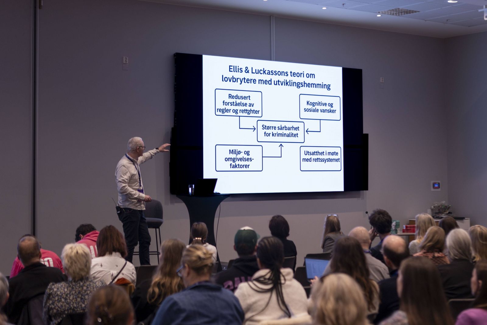 Bildet viser en mann som står på en konferansescene og peker på et lysbilde. publikum i forgrunnen.