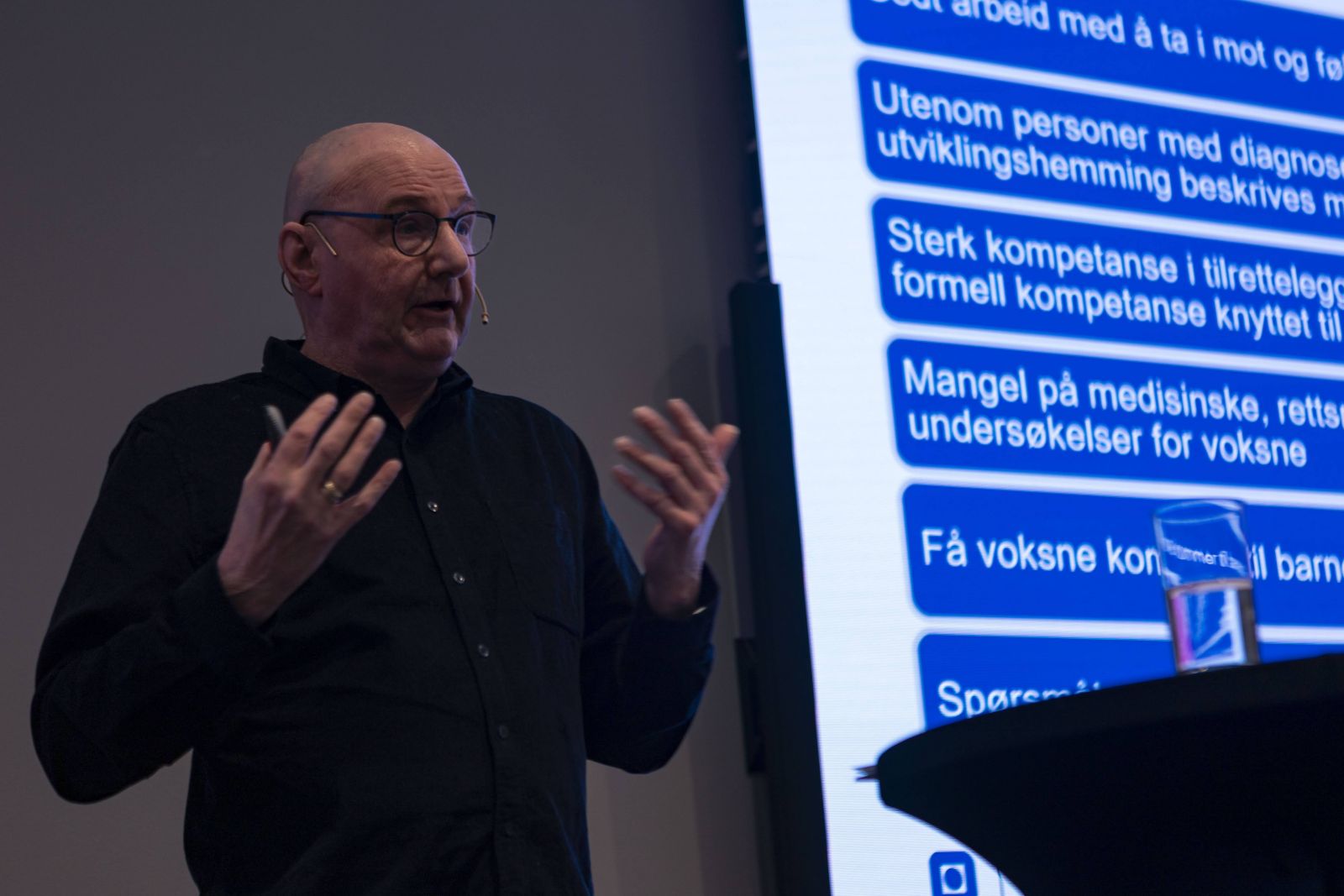 Bildet viser en mann som står på en scene og snakker. lysbile i bakgrunnen.