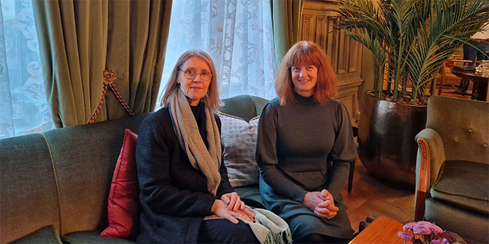 Bilde av to damer som sitter i en grønn sofa. De ser mot fotografen og smiler.