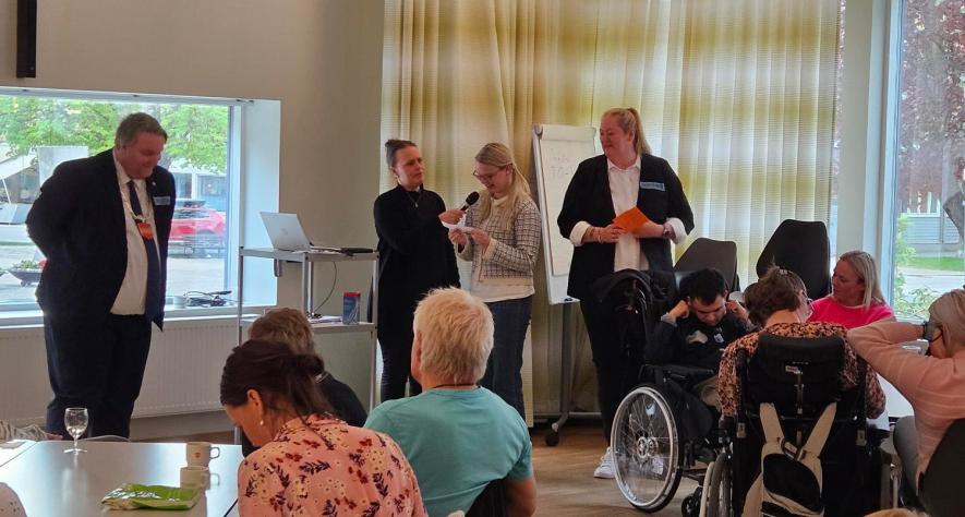 bildet viser en sal med mennesker og i bakgrunnen er scenen der noen står og holder en mikrofon