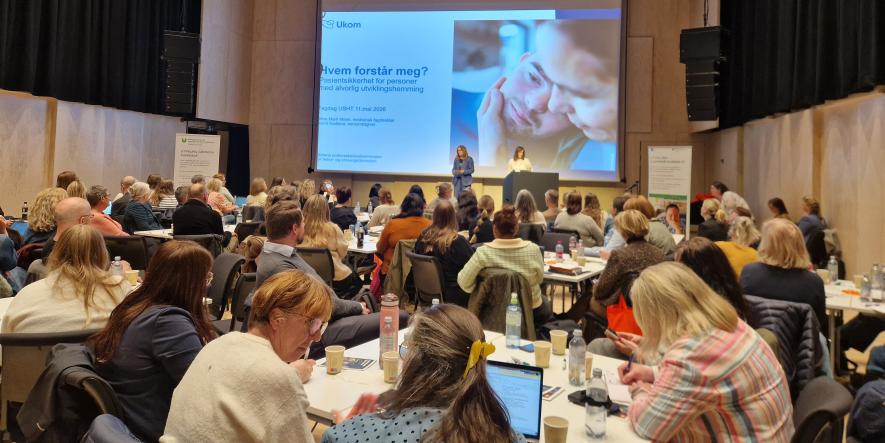 Bilde viser konferanse med deltakere som ser på Powerpointfremlegg på en scene