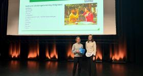 Bildet viser to personer som står på en scene med et lerret i bakgrunnen. På lerretet står "Velkommen til samling".