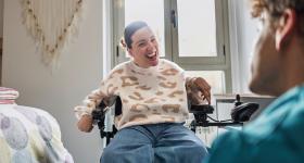 Bilde av en som sitter i rullestol, og smiler mot en som sitter med ryggem mot kameraet. SKjermbilde av forsiden 