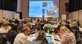 Bilde viser konferanse med deltakere som ser på Powerpointfremlegg på en scene